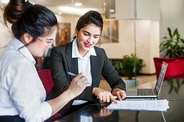 Two women are sitting at a table and one is looking at the paper.